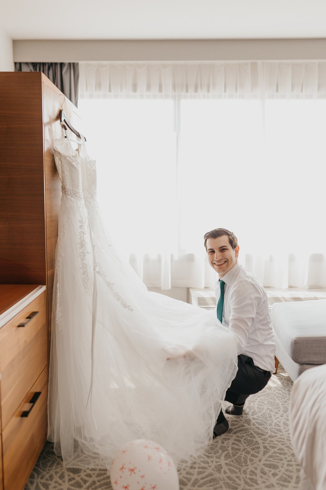 Greg holding Chelsie's wedding dress in a bright hotel room